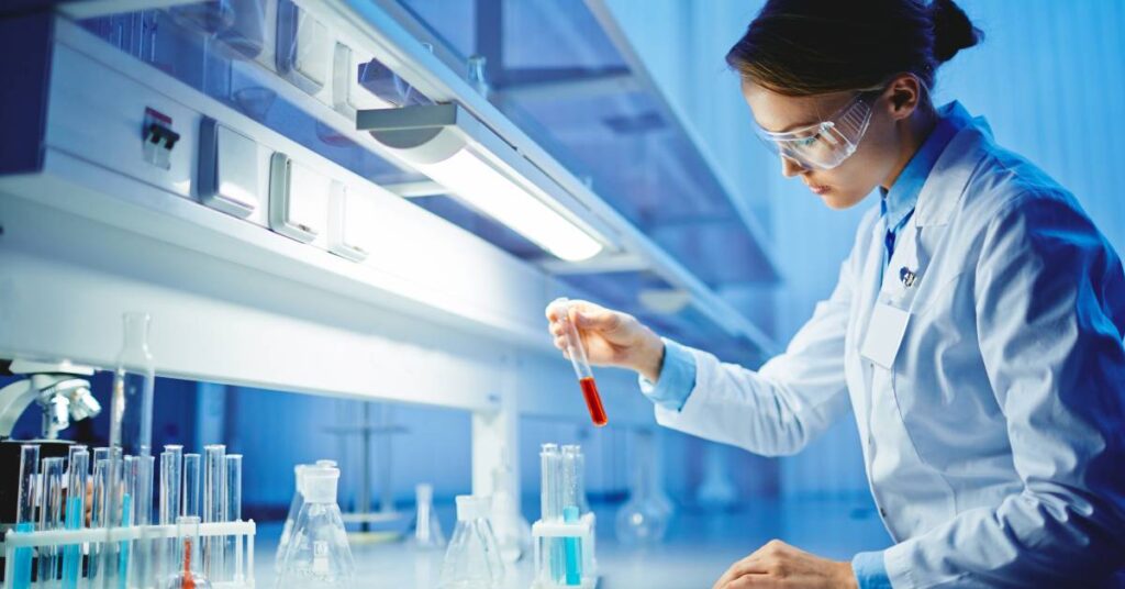 A female scientist standing in a laboratory examining test tubes and taking notes. She is wearing goggles and a lab coat.