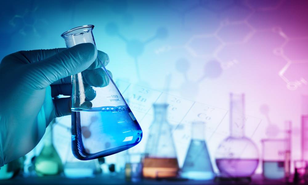 A scientist is wearing gloves and holding a beaker filled with liquid. There are beakers of various sizes in the background.