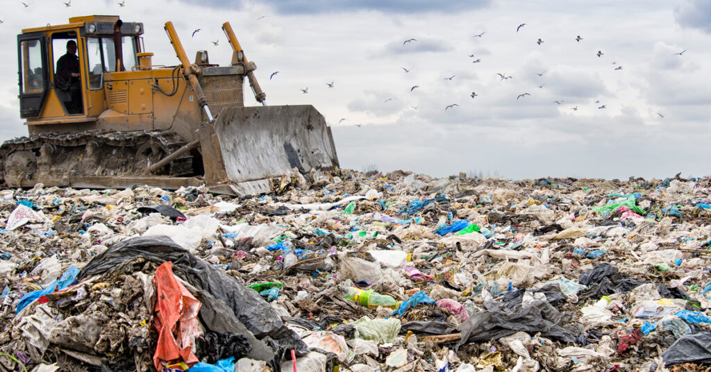 A bulldozer is adding garbage to a large landfill. The sky is cloudy and there are several birds flying above the trash.