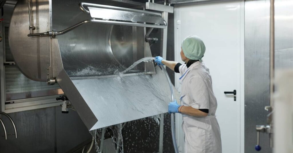 A worker dressed in white protective clothing and a hair net is spraying water on a large piece of stainless steel equipment.