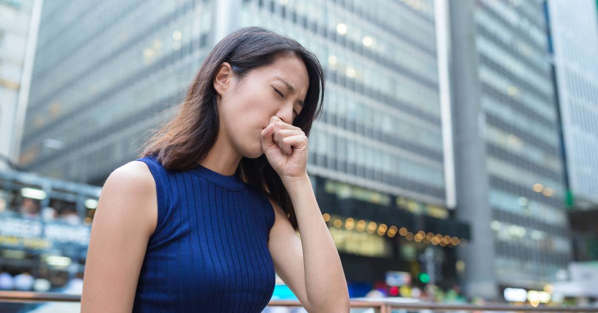 A woman is wearing a blue tank top and standing outside in an urban environment. She is coughing into her hand.