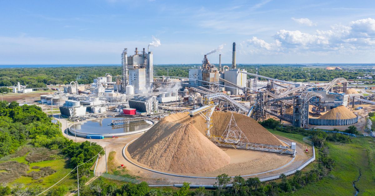The exterior of a large paper mill on a sunny day. There are several chimneys releasing smoke into the air.