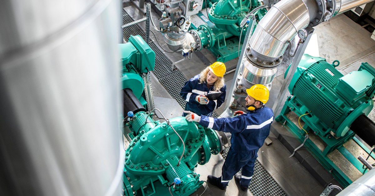 Two workers dressed in protective equipment and hard hats. They are inspecting large gas pipes and generators.
