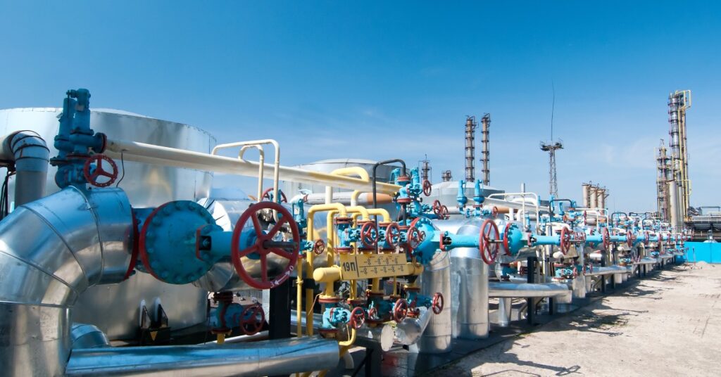 A close-up of several stainless steel gas valves with red and blue components at an outdoor oil refinery.