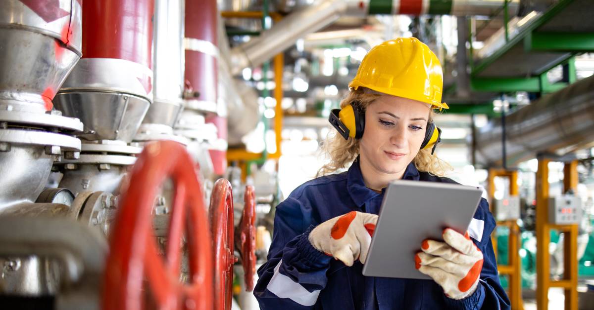 A woman wearing a blue uniform, a yellow hard hat, gloves, and earmuffs is looking at a tablet in a refinery.