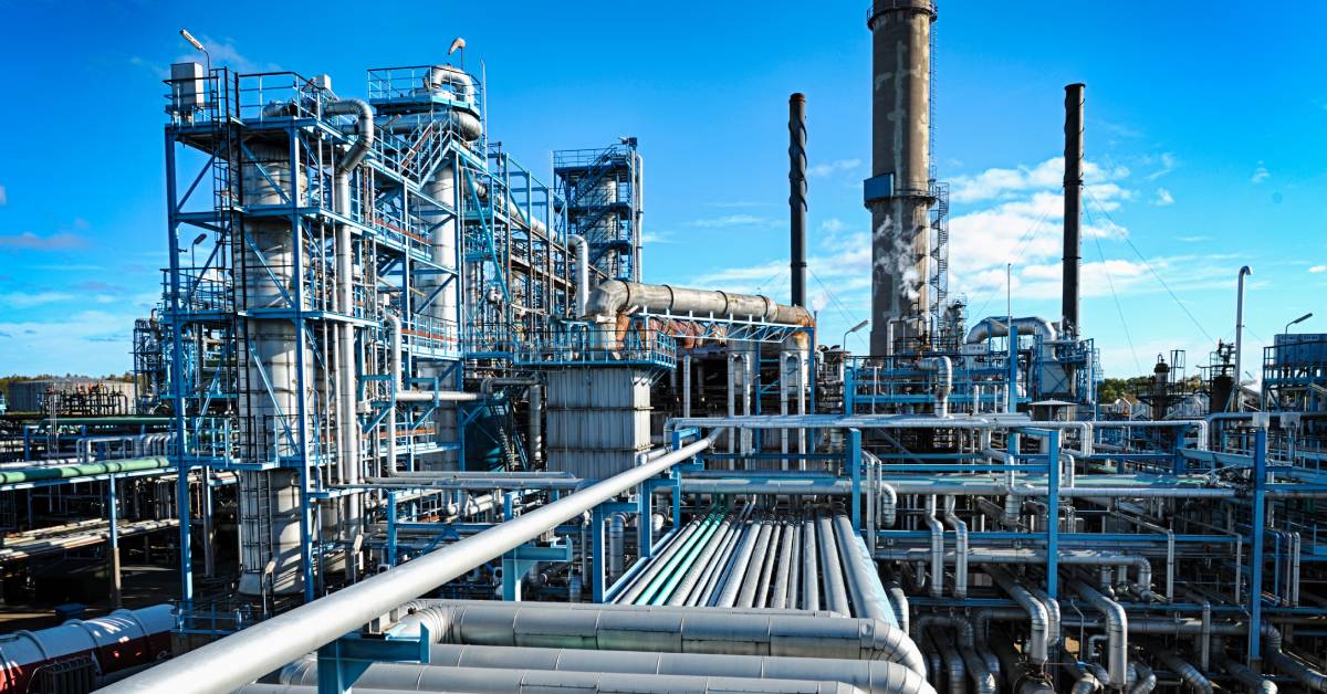 An oil and gas refinery consists of straight and curved metal pipes and tall chimneys. The weather is sunny.