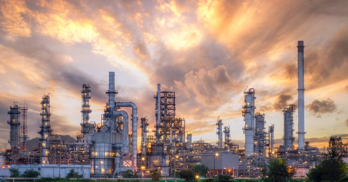 A large oil and gas refinery with multiple buildings and tall chimneys. The sky above the refinery is golden.