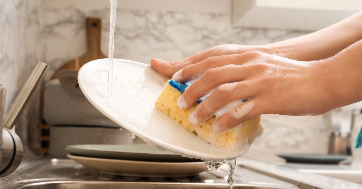 A close-up of a person using soap, water, and a yellow and blue sponge to wash a white plate in a sink.