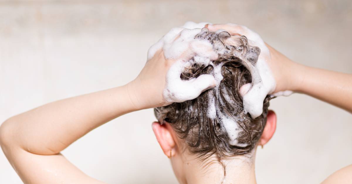 A close-up of a person using both hands to wash their hair. Their hair is filled with white soap suds from the shampoo.