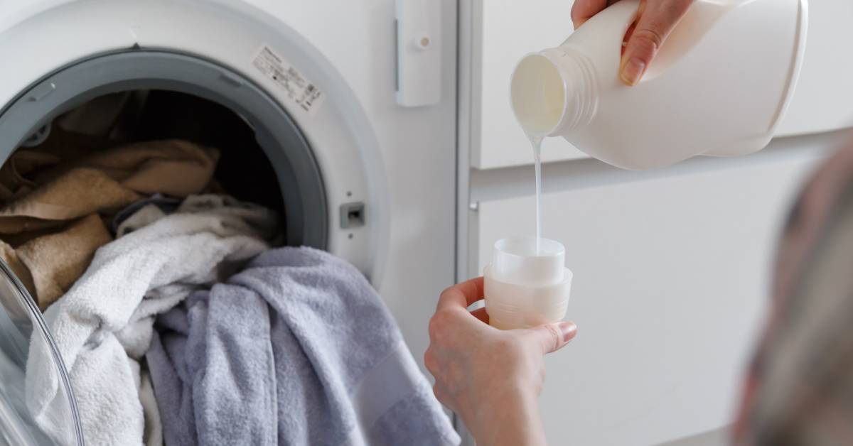 An individual standing in front of an open washing machine and pouring laundry detergent into the cap of a white bottle.