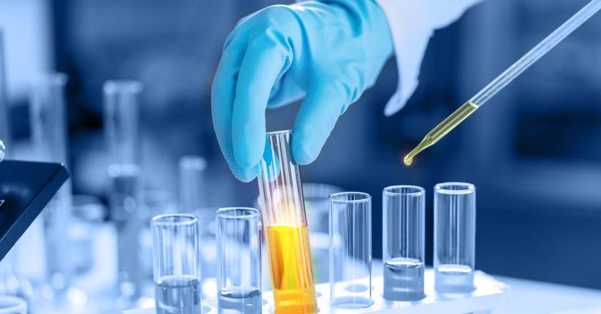 An individual wearing a white coat and blue gloves is holding a glass test tube filled with yellow liquid.