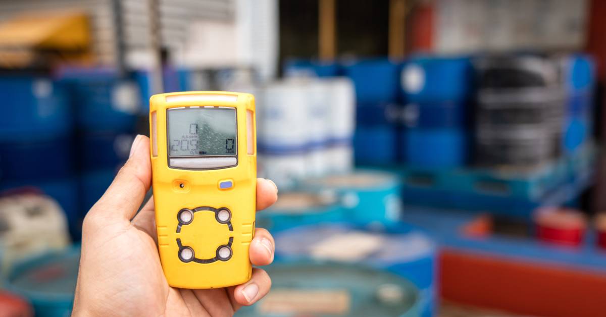An individual is holding a yellow portable gas detector. There are blurred chemical barrels in the background.