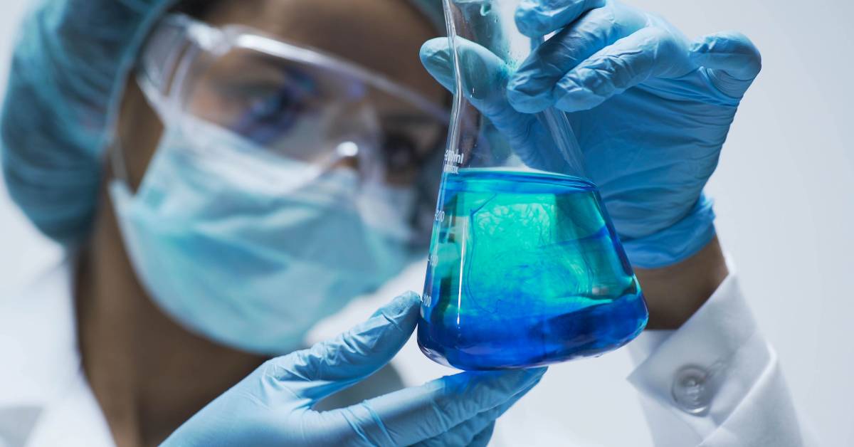 A woman wearing a hair net, protective glasses, a mask, and gloves is holding an Erlenmeyer flask filled with blue liquid.