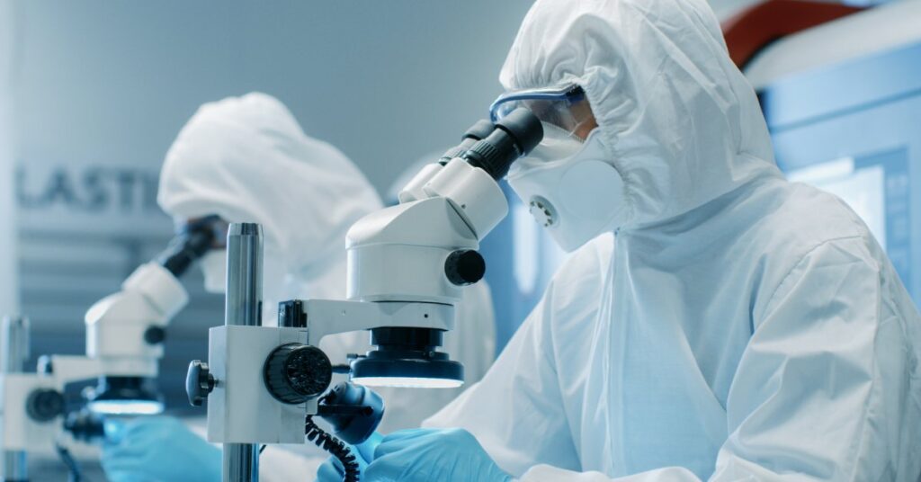 Two individuals wearing white protective suits, blue gloves, and safety glasses are looking into microscopes.