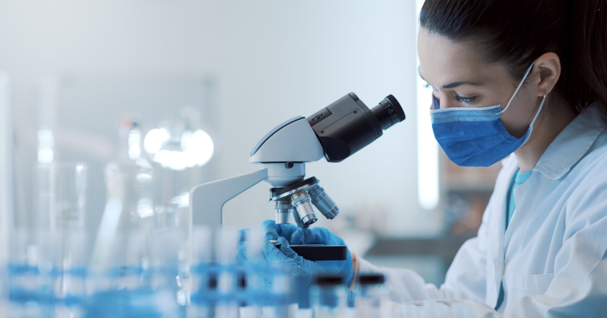A woman is wearing a white coat, blue gloves, and a blue mask. She is analyzing samples under a microscope.