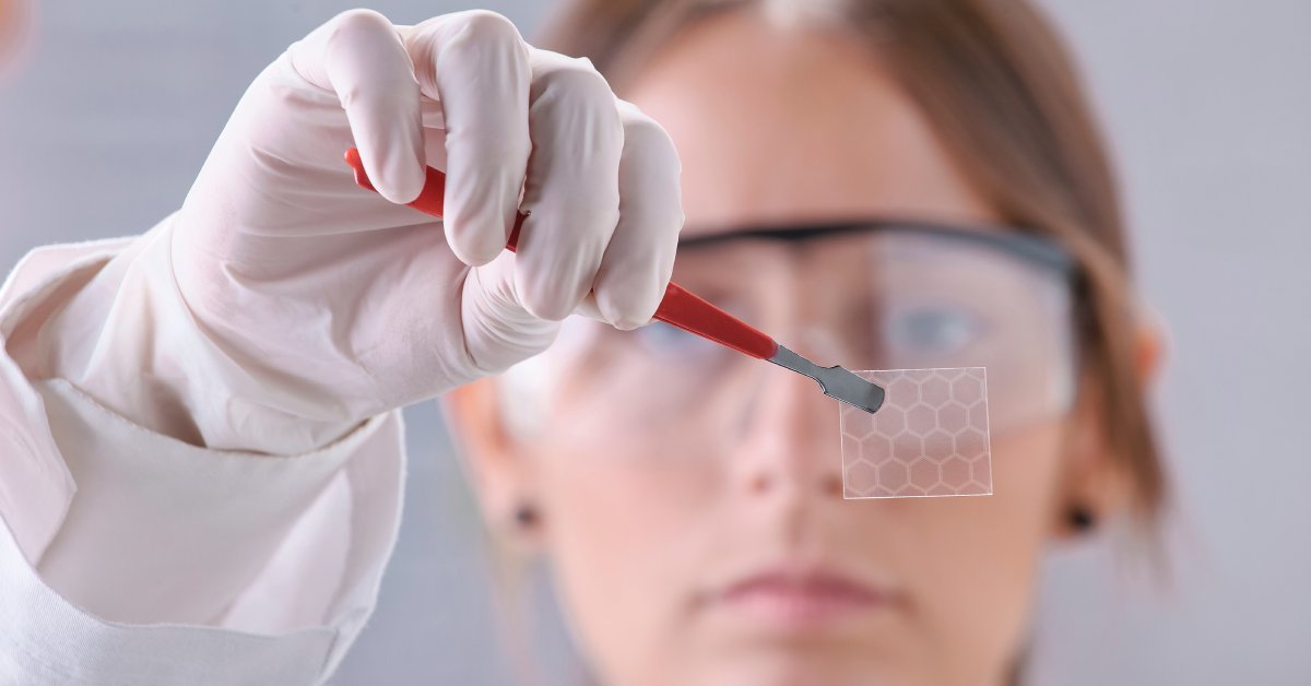 A woman is wearing white gloves and safety glasses. She is holding a small hexagonal sheet with red tweezers.