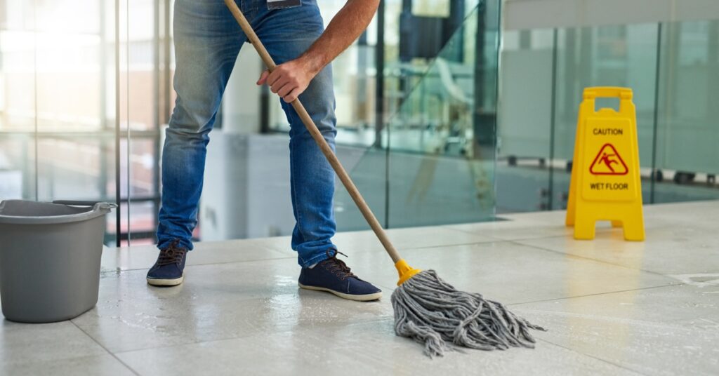 A man wearing jeans is mopping the tile floor of a building next to a yellow sign that says "Caution Wet Floor."