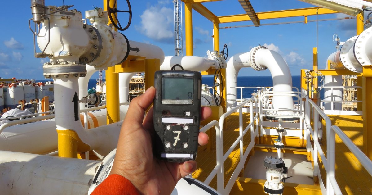 A person's hand holding a yellow hydrogen sulfide gas detector out toward an offshore oil and gas processing platform.