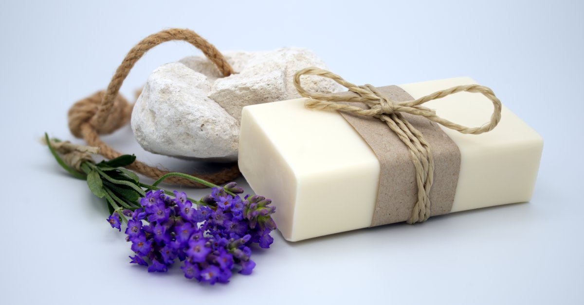 A bar of light-colored soap made from natural ingredients. A bundle of lavender and a pumice stone sit beside it.