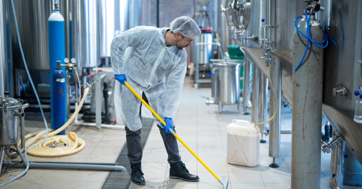 A professional industrial cleaner in a protective uniform is cleaning the floor of a food processing plant.