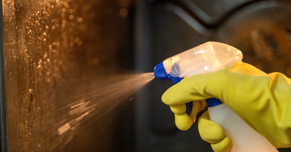 A close-up of a person with plastic yellow cleaning gloves spraying degreaser inside a dirty, greasy oven.