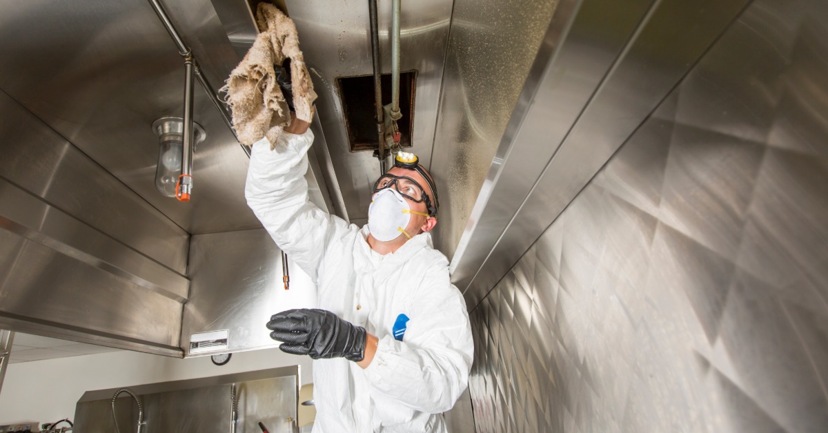 A professional cleaner is wearing a white protective suit, a blue facial mask, and safety goggles while cleaning a kitchen.