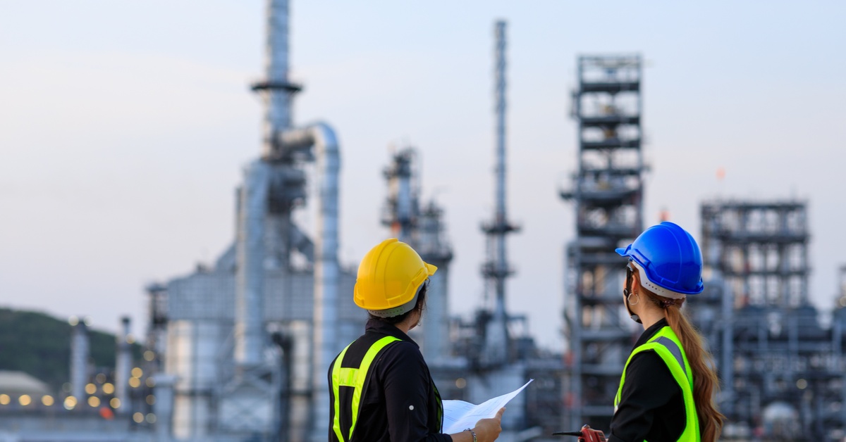 Two female engineers, wearing helmets and holding clipboards, are talking at an industrial gas plant.
