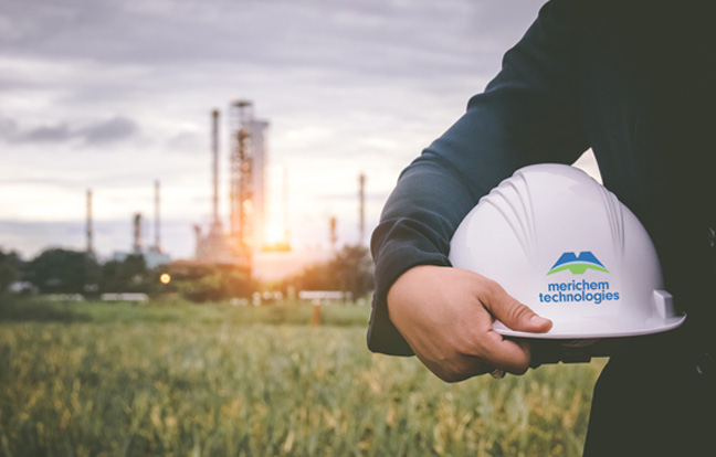 Suited man standing on right side foreground, holding white hard hat, refinery blurred out in the distance in background