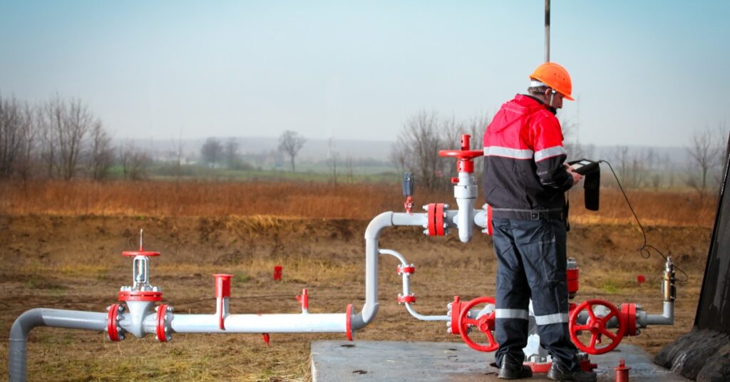 The backview of a gas production operator in a red windbreaker and a hard hat working outside and tracking the gas usage.