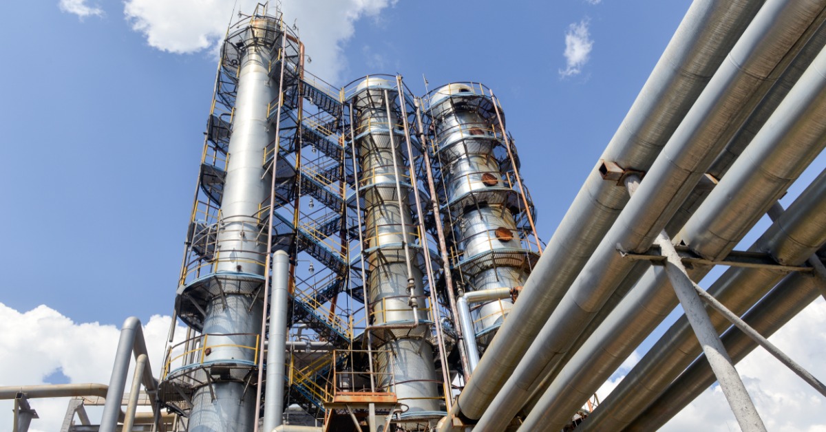Industrial refinery towers and pipelines at a large oil or gas processing plant under a bright blue sky with clouds.