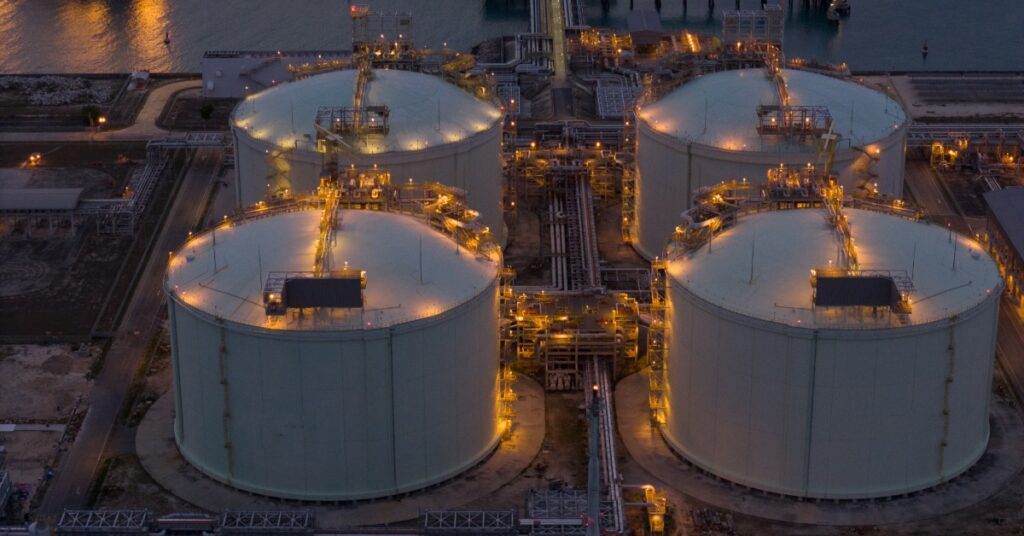 Four large storage tanks with lit walkways and piping at an industrial site near water during evening hours.
