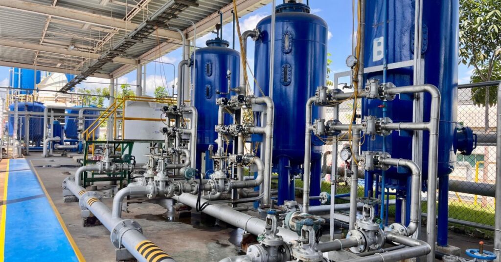 Industrial facility with blue vertical tanks, pipes, and valves arranged in rows under a metal roof structure.