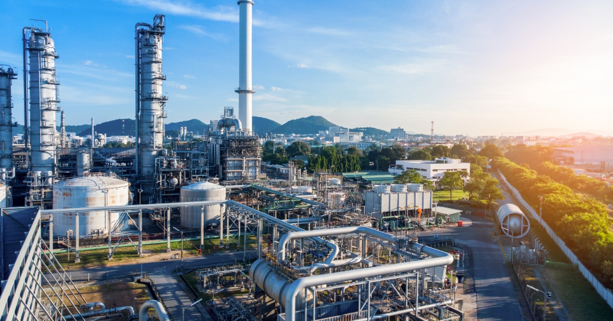 Large refinery complex with metal structures, pipelines, and smokestacks near a city, viewed under clear blue sky.