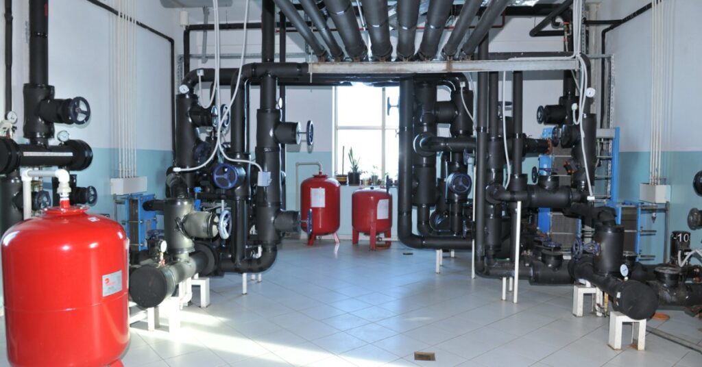 Bright utility room shows large black pipework, handwheel valves, gauges, and red tanks for building systems.