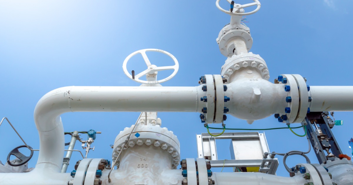 White industrial pipes and handwheel valves connect with bolted flanges at a refinery beneath a clear blue sky.