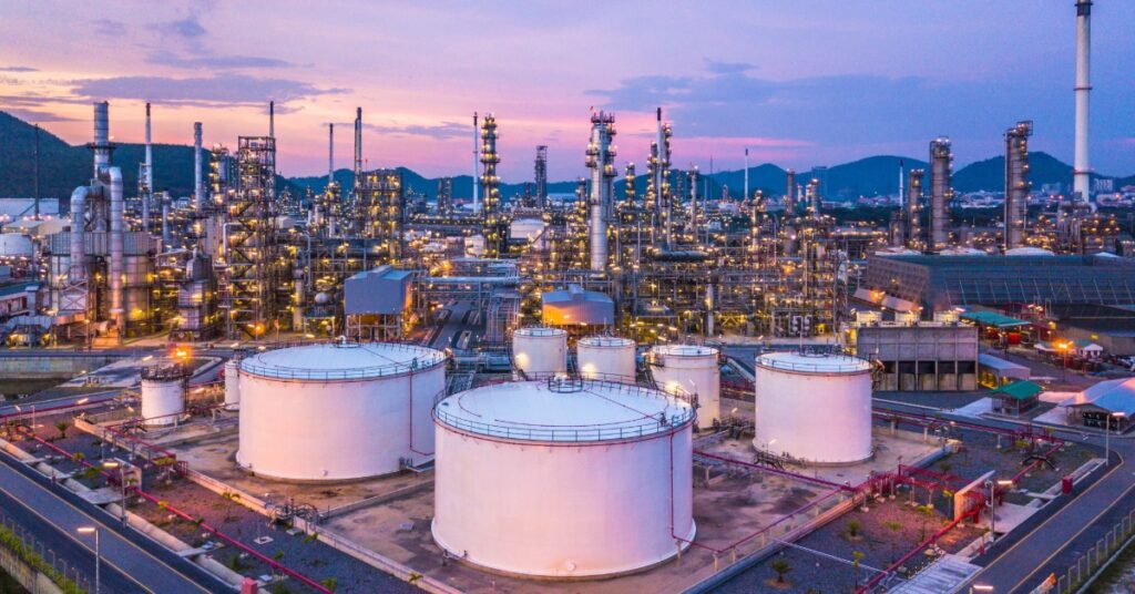 An oil refinery with oil and gasoline chemical tanks and barrels. The sun is setting in the background.