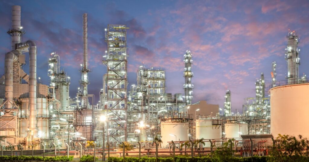 Brightly lit refinery with tall processing towers, storage tanks, and pipework under a colorful dusk sky.