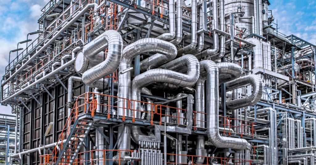 Close view of industrial refinery with insulated pipes, metal platforms, and processing equipment under blue sky.