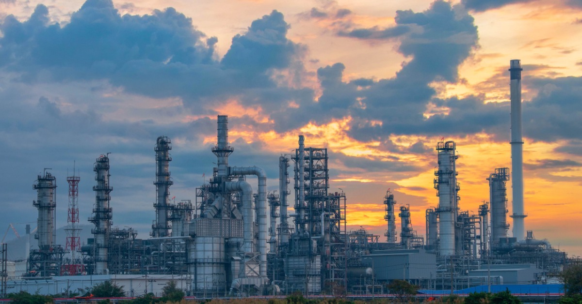 Oil refinery towers and pipes form a dense industrial skyline beneath dramatic clouds glowing orange at sunset.