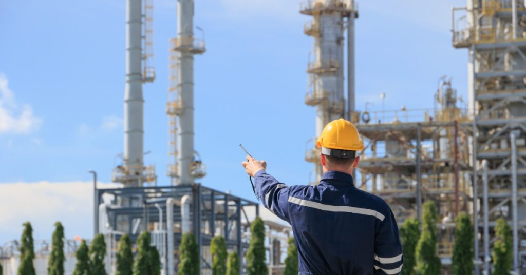 Worker in a yellow hard hat points toward refinery towers and metal structures at an industrial plant under blue sky.