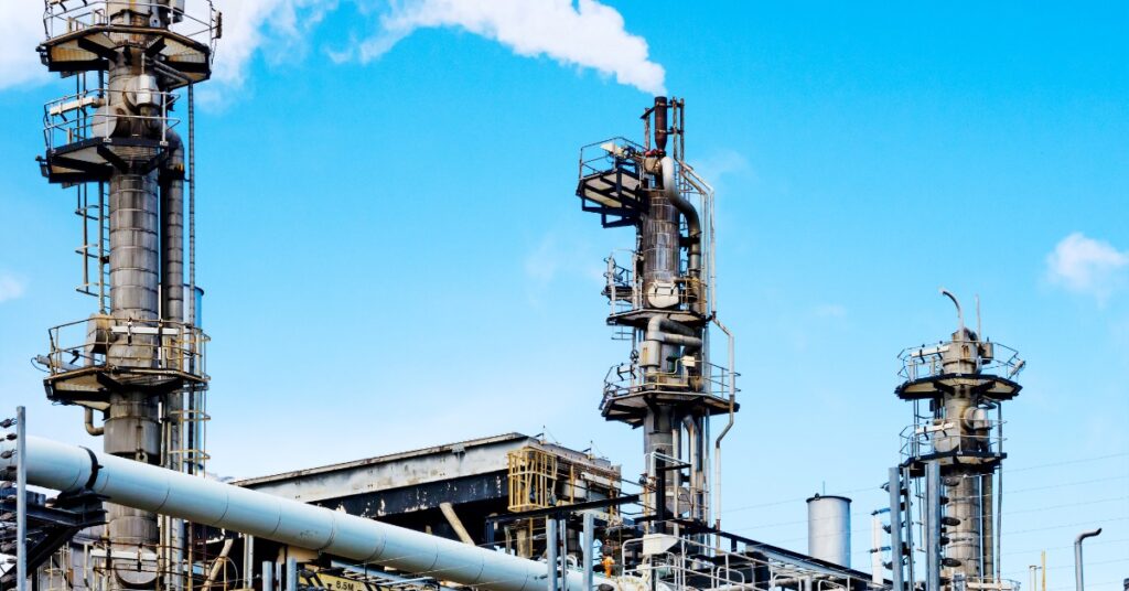 Oil refinery towers, pipes, and metal platforms rise under a bright blue sky with white vapor drifting from a stack.