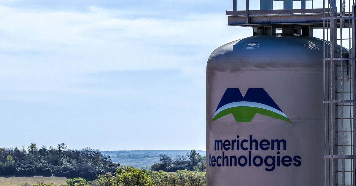Vertical SULFURTRAP H2S removal vessel with Merichem Technologies blue and green logo on it against blue sky with tree-lined field visible across horizon